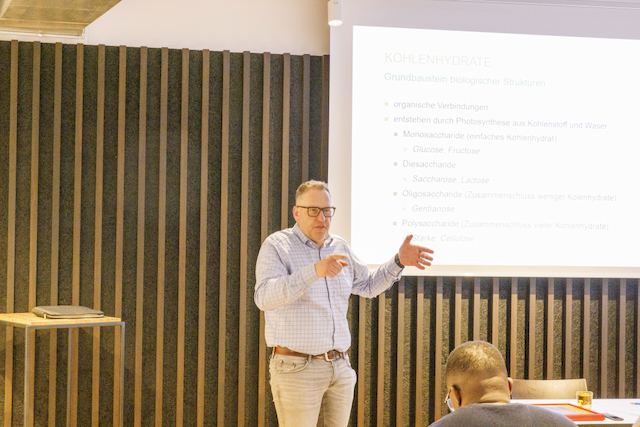 Ein Mann präsentiert beim Macardo Brenn-Kurs vor einer Leinwand mit Text über Kohlenhydrate. Er trägt ein hellblaues Hemd, graue Hose und Brille, gestikuliert mit beiden Händen. Ein Teilnehmer sitzt im Vordergrund.